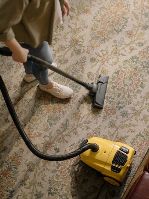 A woman using a yellow vacuum cleaner with a black hose to deep clean a patterned area rug in a living room. The rug features a floral design with muted tones, and the room has wooden flooring and a r